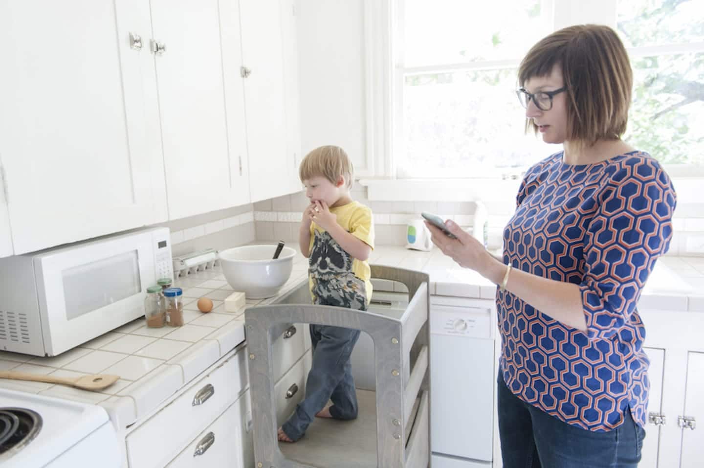 woman looking at her phone and child looking at a bowl in the kitchen