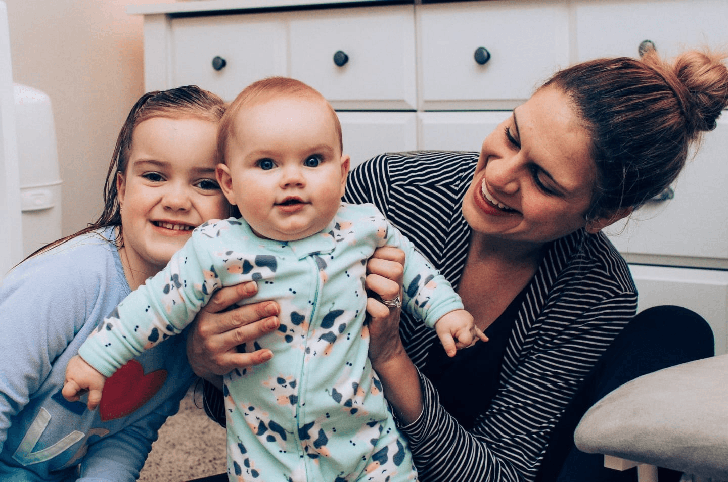 mom holding baby while toddler smiles beside her