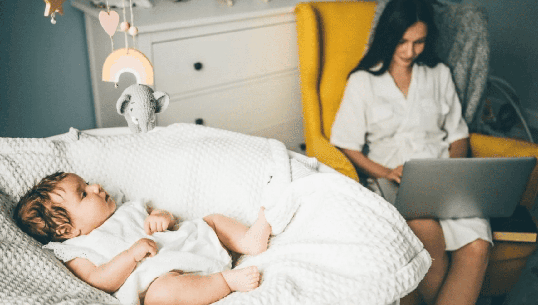 mom sitting in glider working on a computer while a baby lays in a carrier