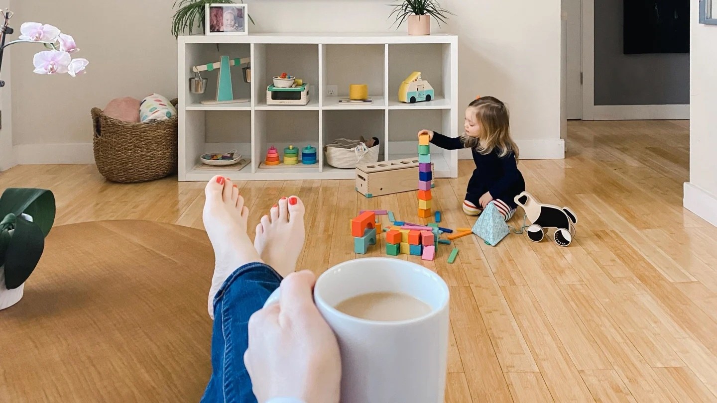 mom holding cup of coffee with her feet up while her daughter plays with blocks in front of her