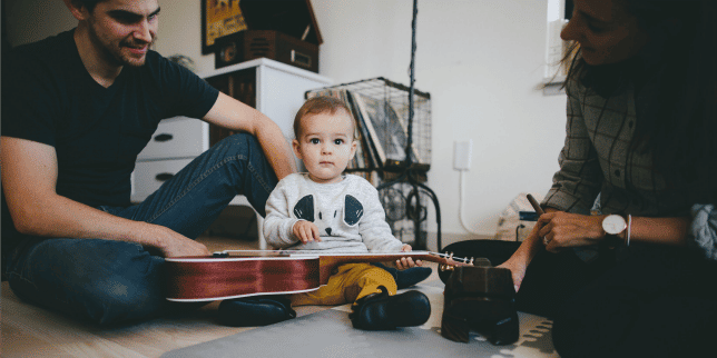 parents smiling as baby sits on the floor and plucks at a guitar, one of Motherly's favorite learning activities for baby