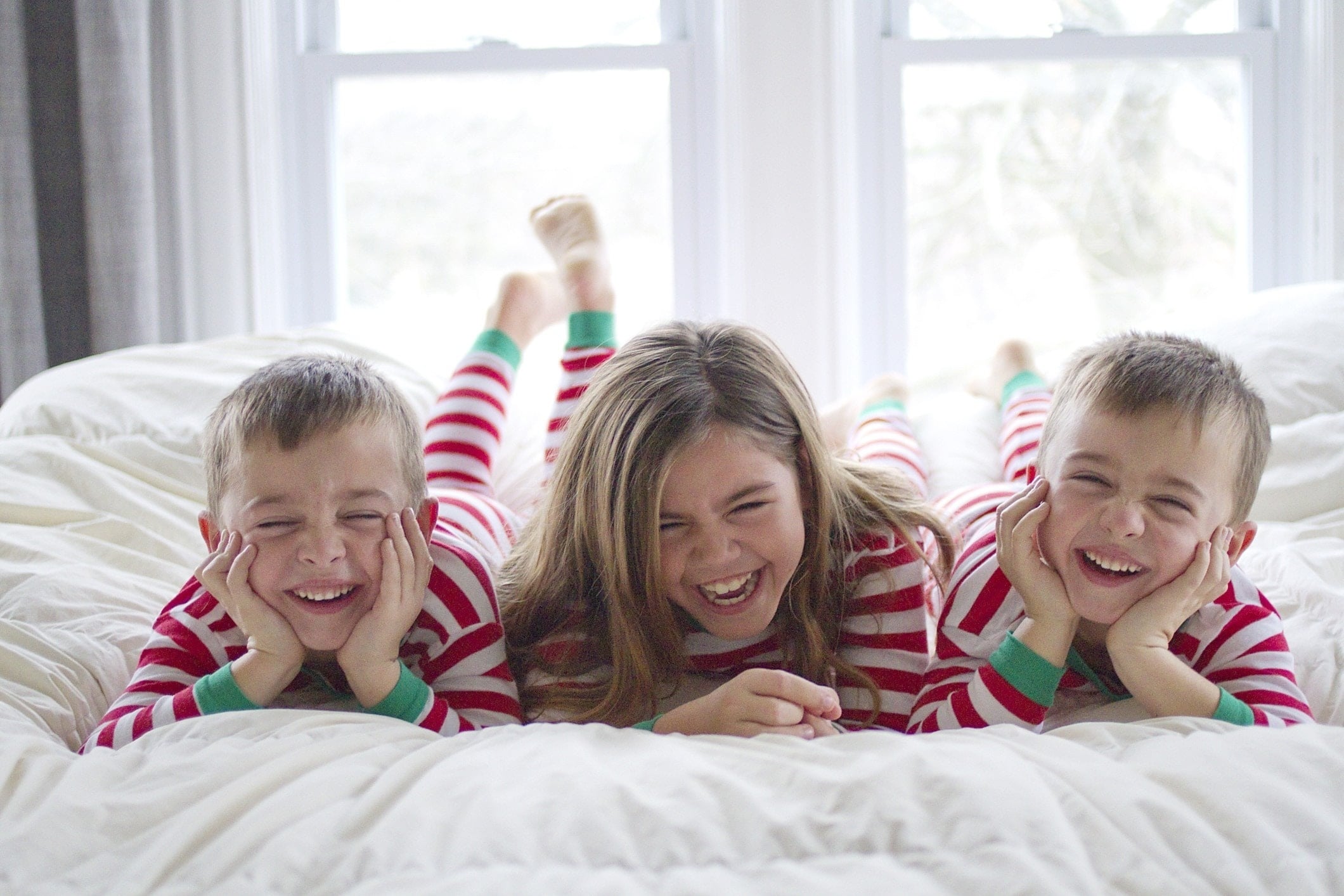 three kids in Christmas pajamas giggle on the bed - research shows giving kids fewer toys at Christmas contributes to happiness