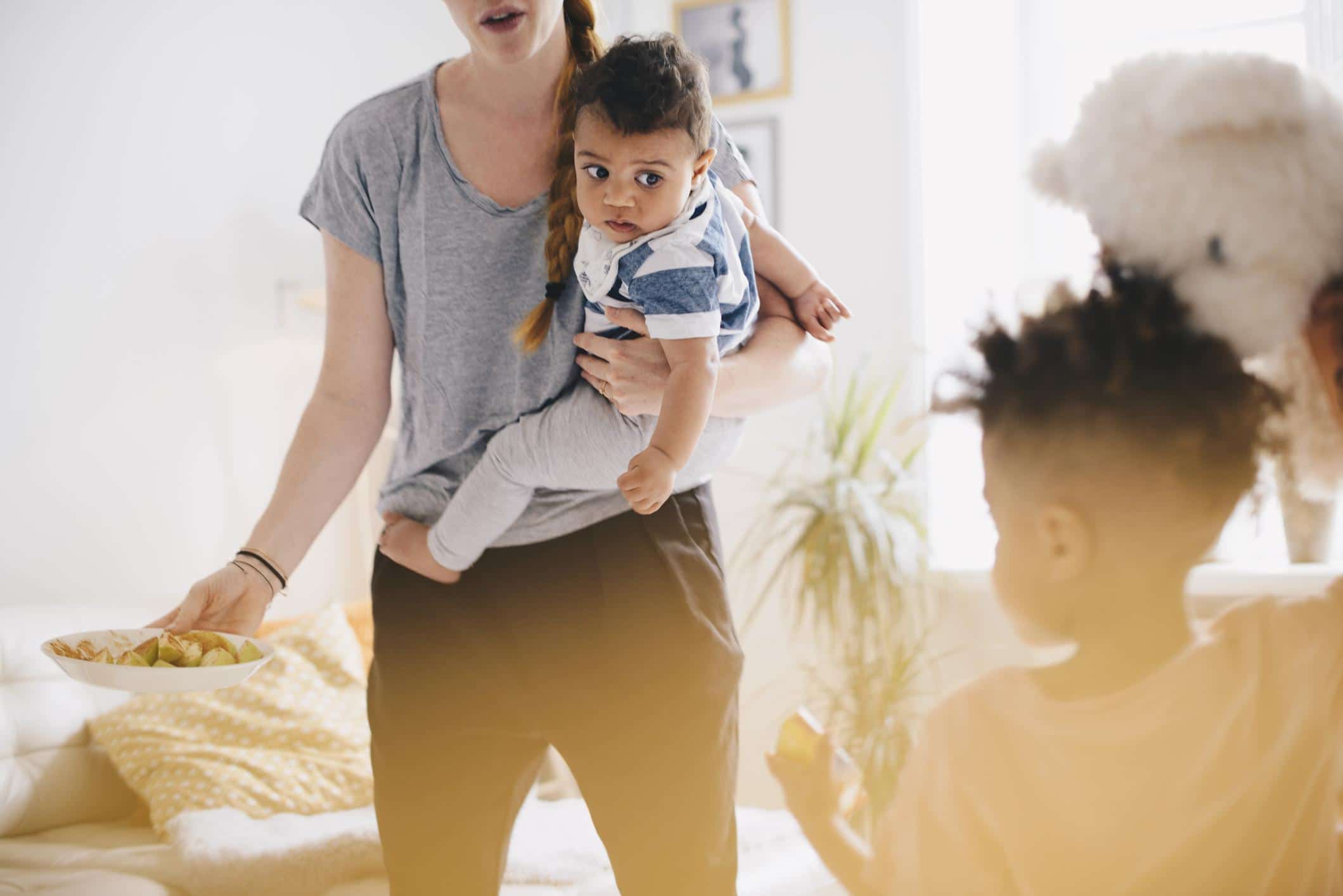 mom holding baby while toddler plays beside her