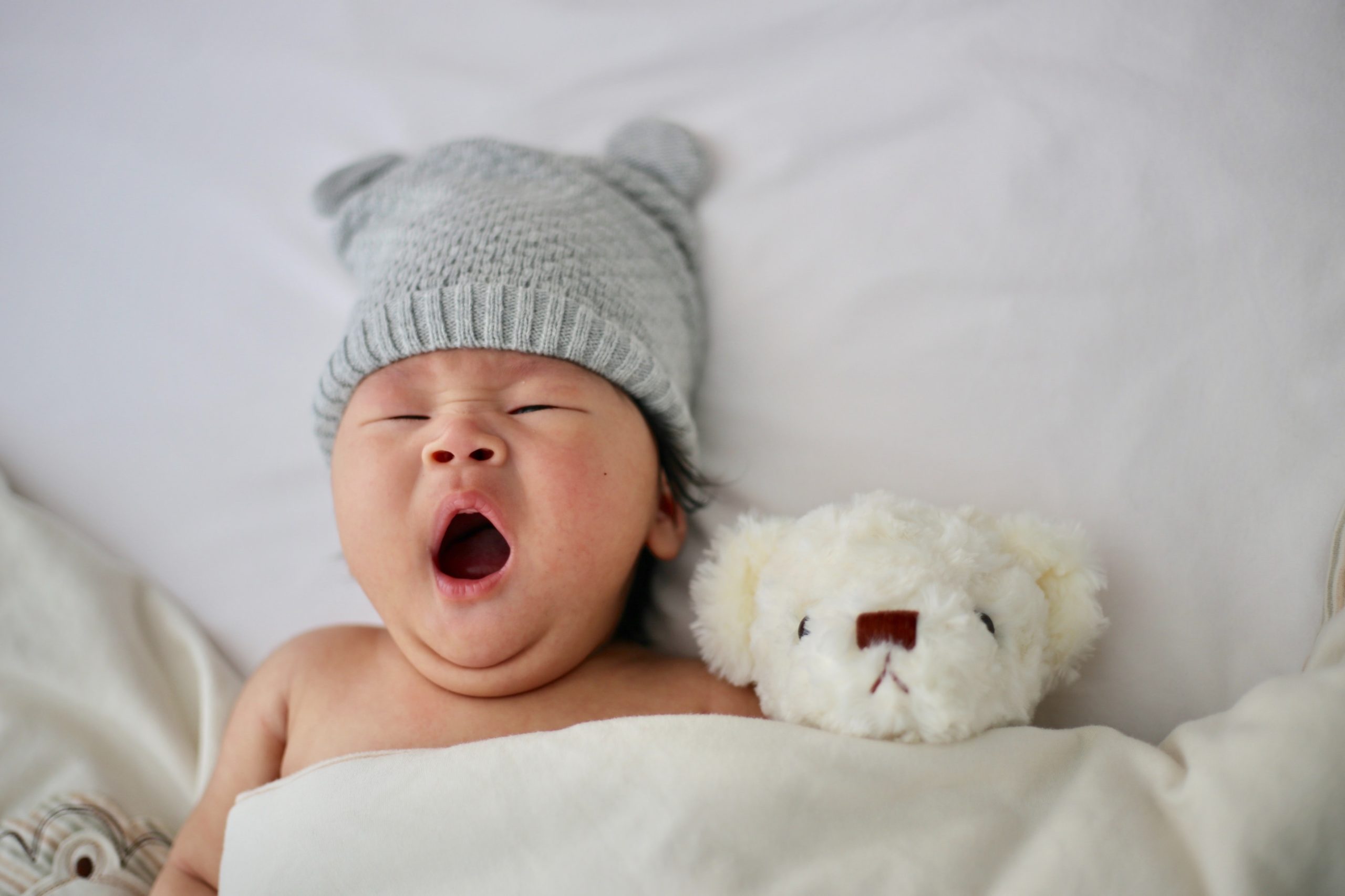 baby yawning wearing a cap and sleeping next to a stuffed animal