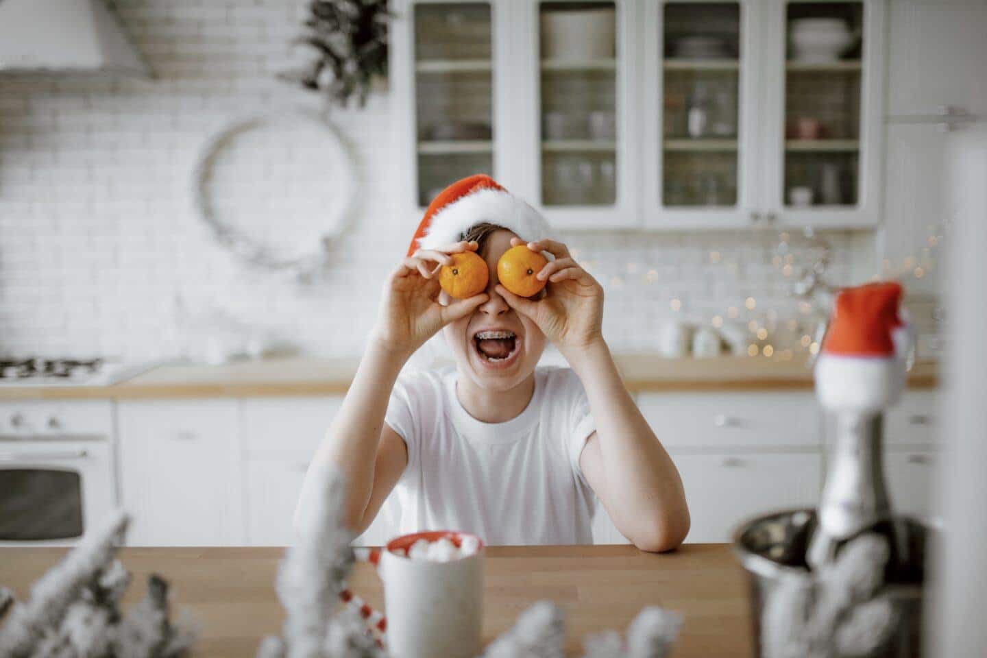 Kid wearing a Santa hat holding oranges up to his eyes - best christmas jokes for kids