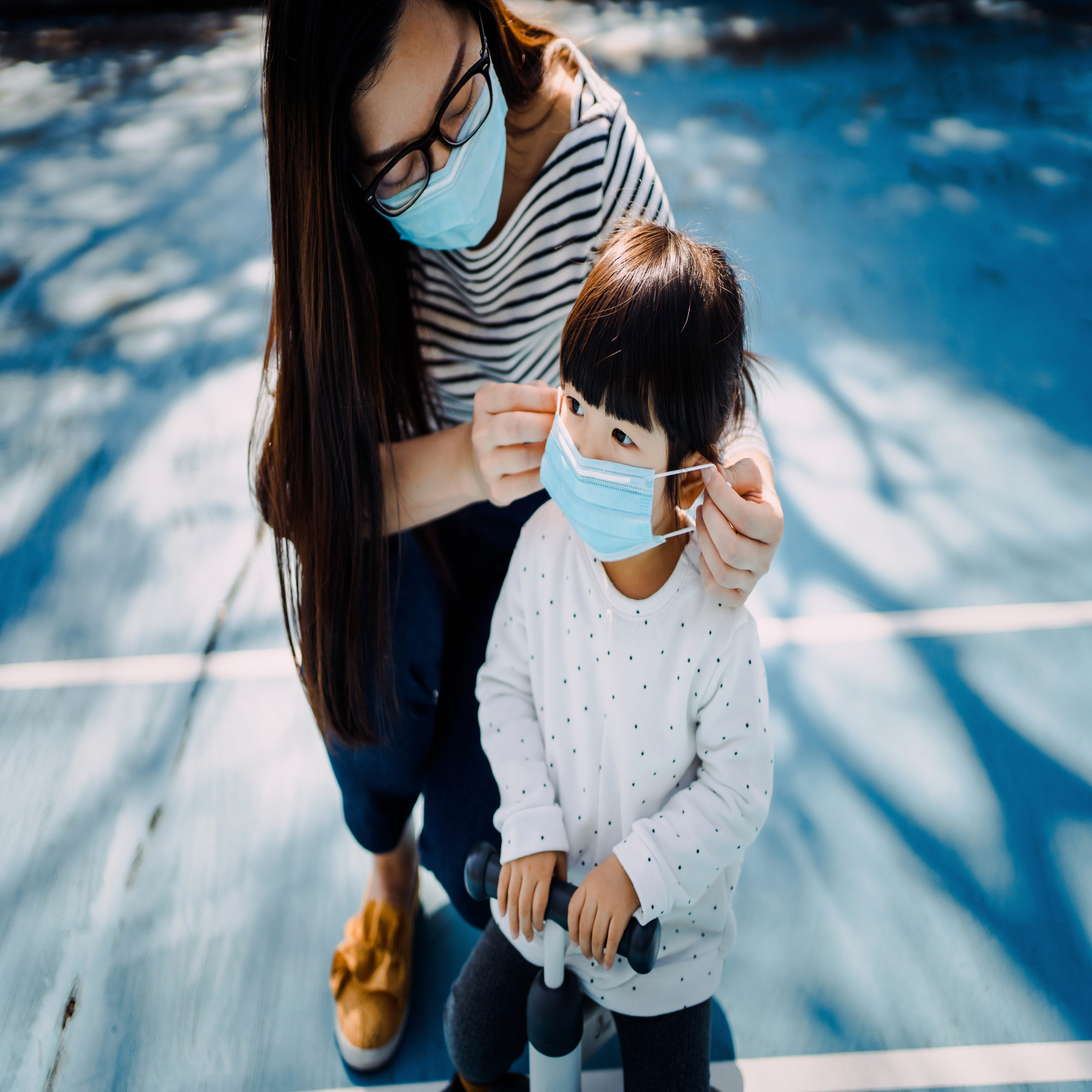 Kids face masks; Young mother putting on surgical mask for little daughter in the playground to prevent the spread of cold and flu and viruses
