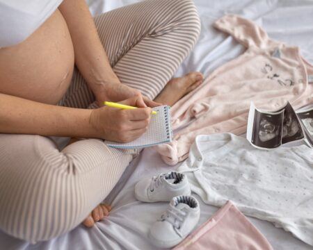 top view of pregnant woman in the bed preparing baby clothes at home and writing a to do list and t20 YN83BE