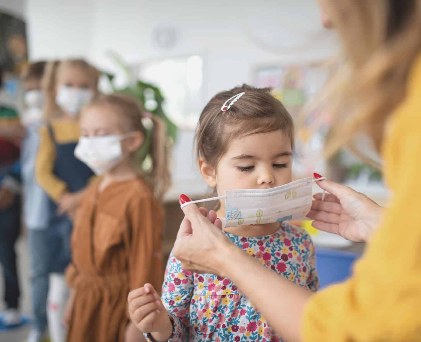 Preschool girl gets help from teacher to put on covid face mask