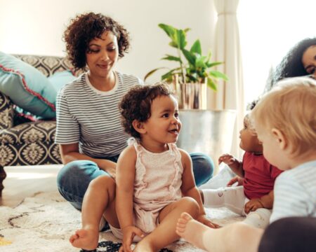 Mom friends playing with toddlers at home showing beauty of postpartum care after fourth trimester Motherly