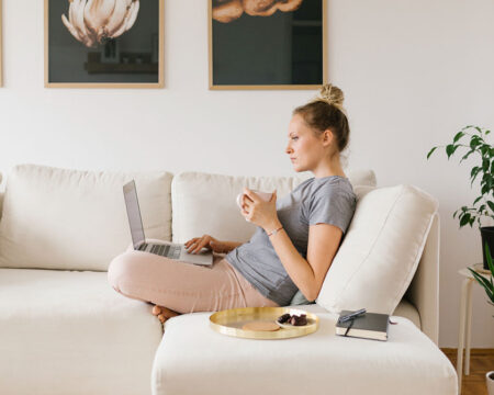 woman sitting on her couch drinking coffee and checking her laptop