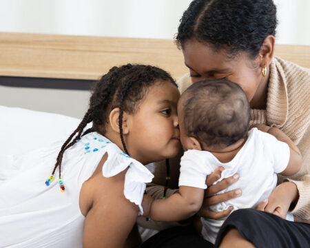 mom on the bed with her daughter and baby giving kisses 1 Motherly