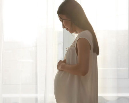 pregnant woman standing near window at home thinking about having a third child Motherly