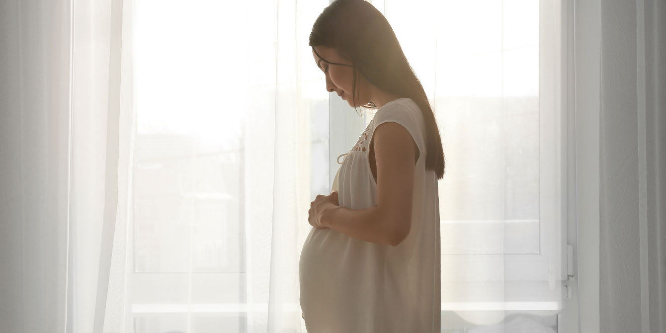 pregnant woman standing near window at home thinking about having a third child