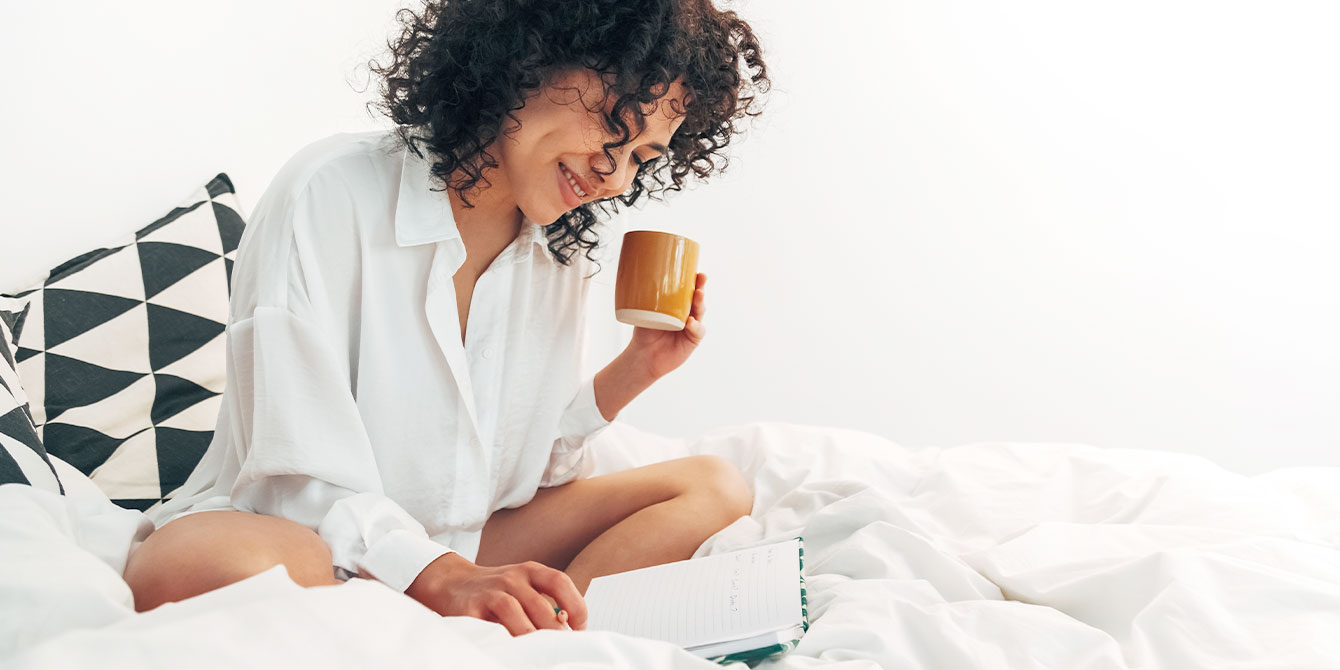 woman in bed drinking coffee and writing in guided journal