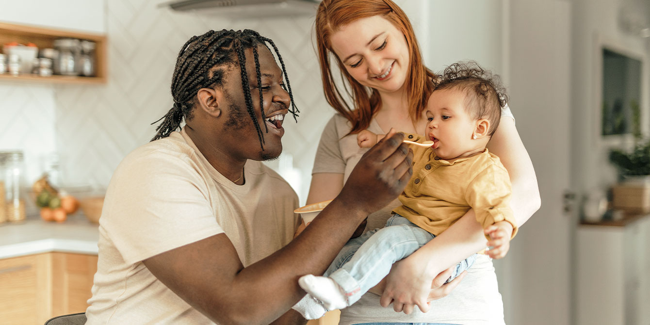 smiling mom holding baby while smiling dad feeds baby baby food